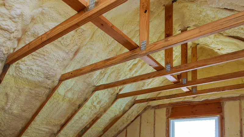 Attic with wooden beams insulated with yellow spray foam covering roof and walls around a window.