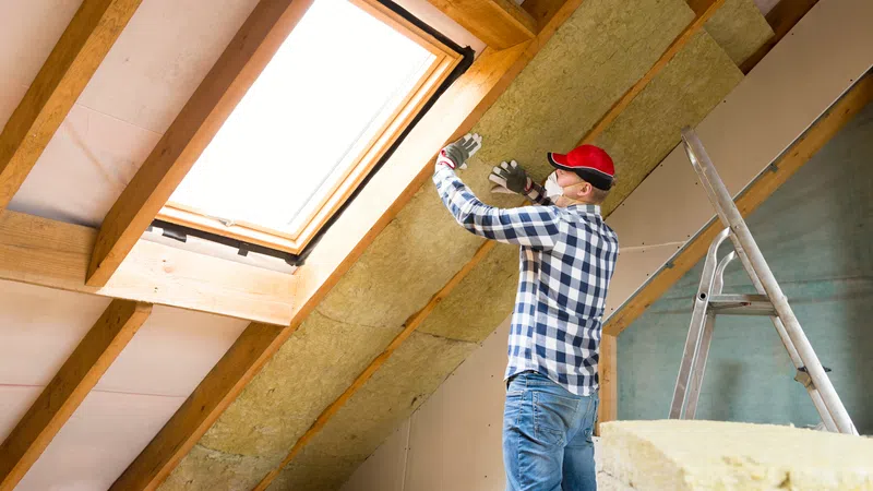 Worker installing insulation panels under a roof skylight wearing safety gear and gloves indoors.