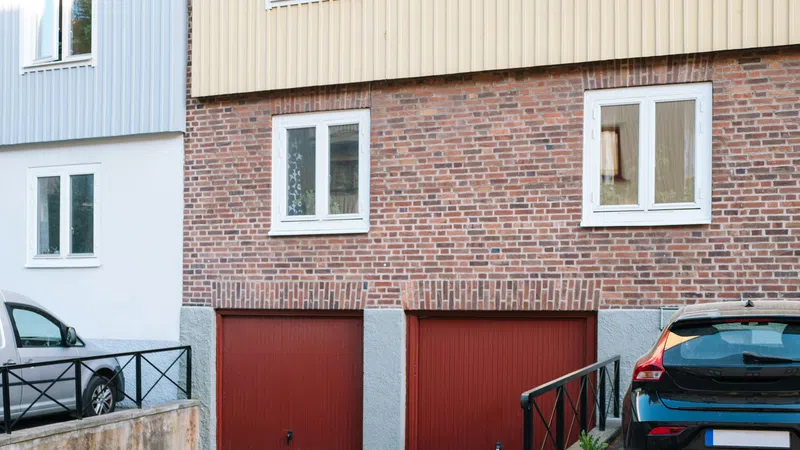 Residential building exterior with brick wall, two red garage doors, and parked black car beside them.