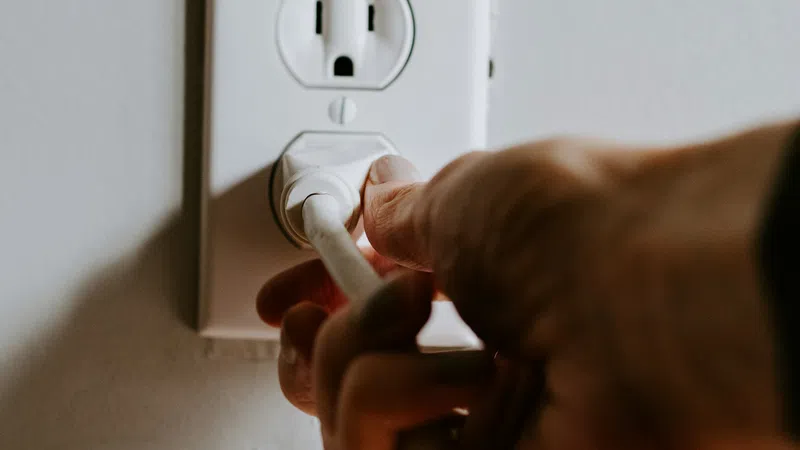 Hand plugging a white electrical cord into a white wall outlet on a light-colored wall.