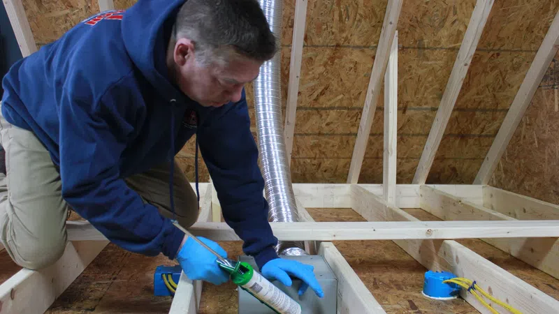 Worker applying sealant around electrical box in attic construction with wooden framing and ductwork visible