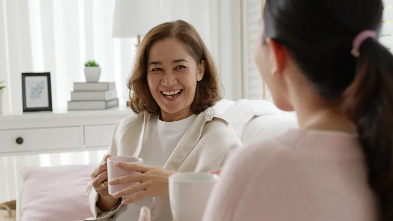 Two friends enjoying coffee and laughter in a bright, cozy living room.