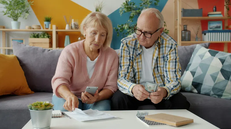 Elderly couple sitting on a sofa reviewing bills and counting cash in a colorful living room