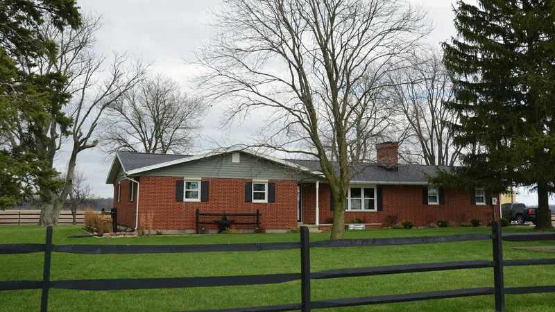 One-story red brick house with black shutters behind black wooden fence on green lawn under cloudy sky