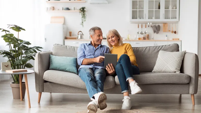 Smiling senior couple sitting on sofa using tablet in bright modern living room with cozy decor