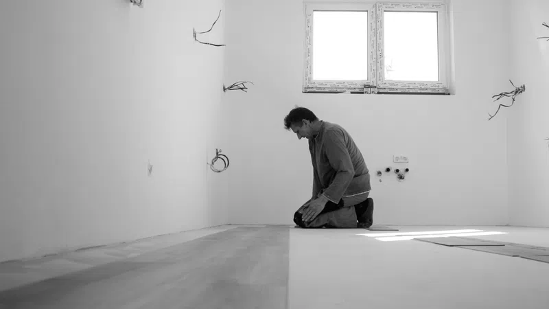Man installing wooden flooring in a bright empty room under renovation with exposed wiring on walls.
