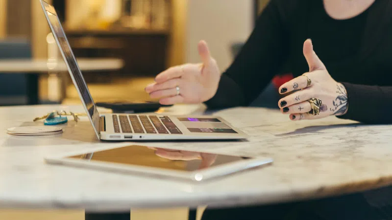 Person with tattooed hands gesturing near a laptop on a marble table in a cozy indoor setting.