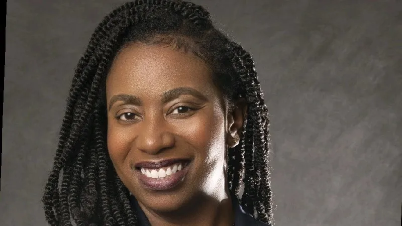 Smiling woman with long curly hair wearing a navy blazer and polka dot blouse on gray background