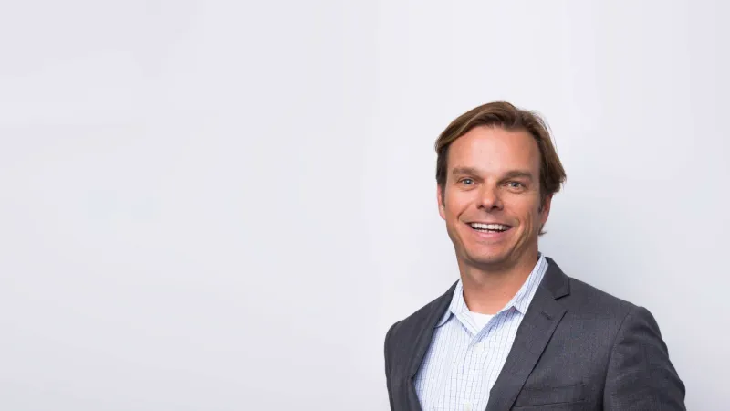 Smiling man in gray suit and checkered shirt standing with hands in pockets against light background