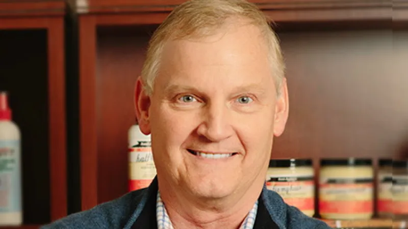 Smiling middle-aged man in blue vest and checkered shirt standing with arms crossed in front of shelves with bottles and jars