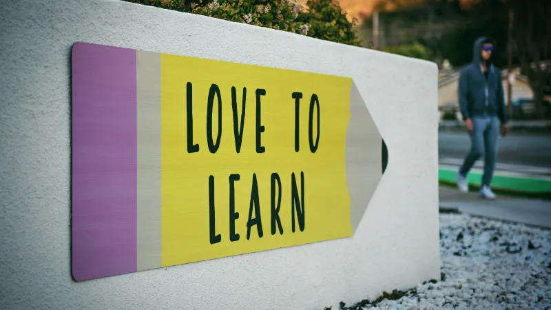 Large pencil-shaped sign with the text 'LOVE TO LEARN' mounted on a white wall near a street.