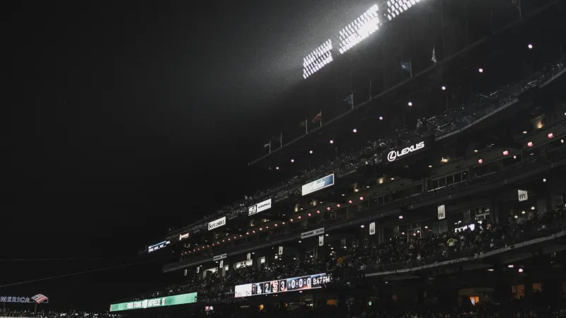 Nighttime view of a crowded baseball stadium with bright floodlights illuminating the field and spectators.