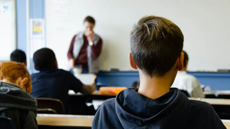 Students seated in a classroom listening to a teacher standing near a whiteboard during a lesson.