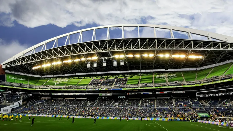 Wide view of a soccer stadium with green seats, players warming up on the field, and a cloudy sky above