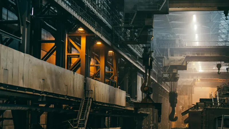 Interior of a large industrial factory with metal structures, machinery, and overhead cranes under dim lighting.