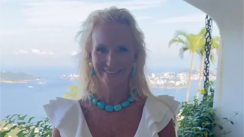 Woman in a white ruffled dress with turquoise jewelry standing on a balcony overlooking a coastal landscape.