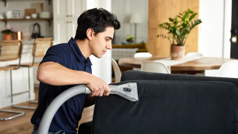a male Zerorez cleaning technician using a handheld upholstery cleaning tool to steam clean a dark microfiber couch cushion in a living room using Zr Water®