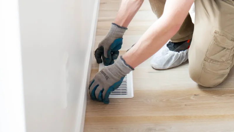 a man squatting on a wood floor lifting up a house heating vent with urine smell