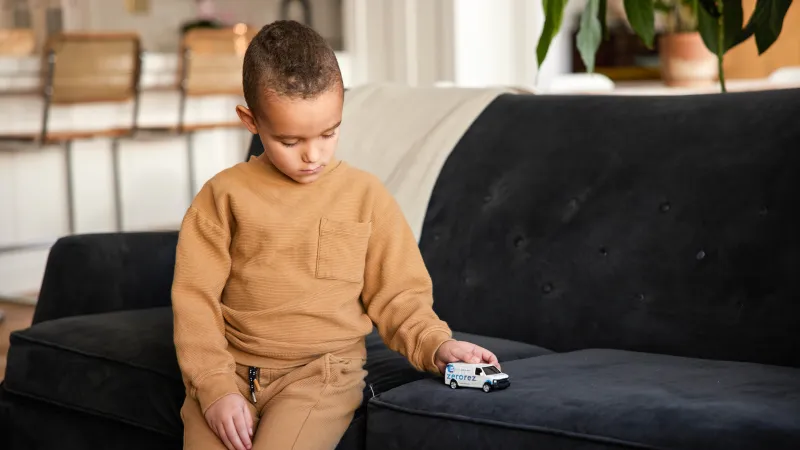 a little boy in a tan sweat suit sitting on a suede couch playing with a small Zerorez toy van