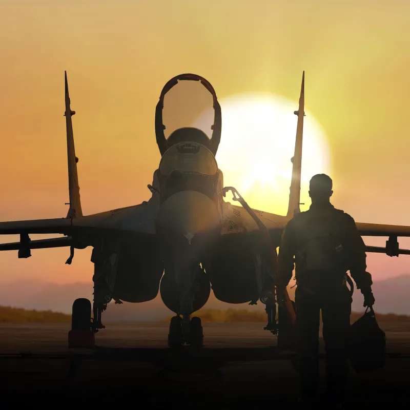 Silhouette of fighter jet and pilot walking on runway during sunset with sun low on horizon.