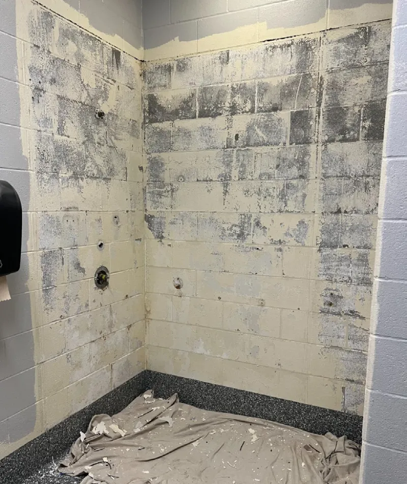 Corner of room with peeling paint on cinder block walls and drop cloth on floor, labeled Before renovation.