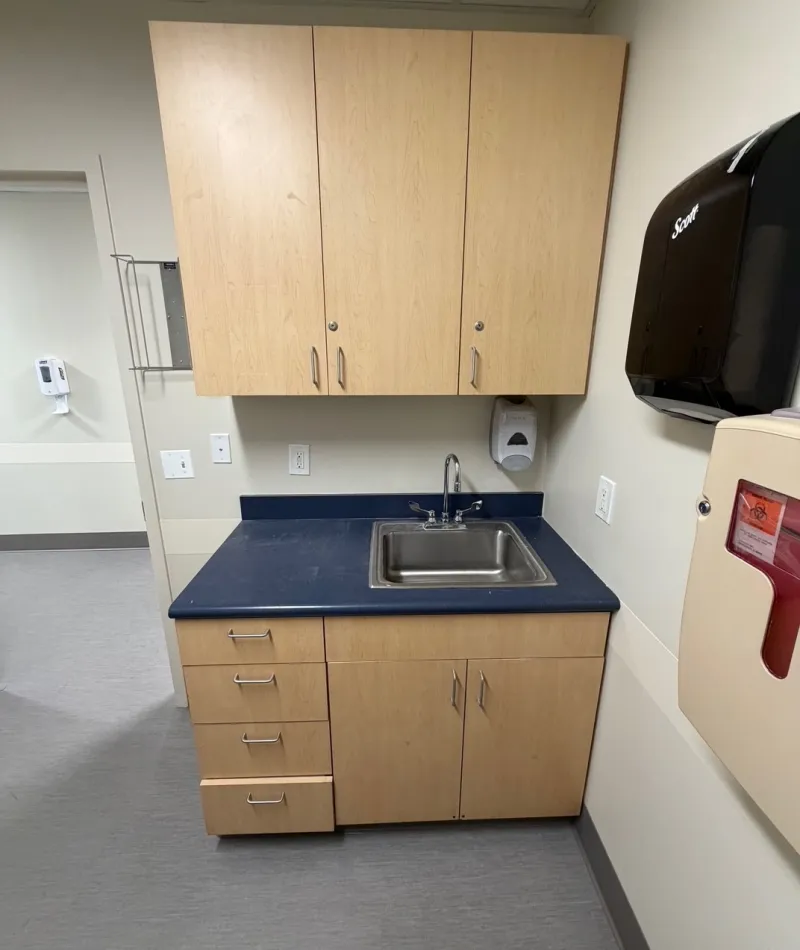 Medical office sink area with wood cabinets, blue countertop, soap dispenser, and paper towel dispenser on beige walls.