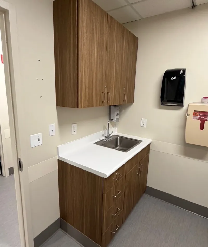 Small medical room sink with wood cabinets, white countertop, hand sanitizer dispenser, and medical waste bin.