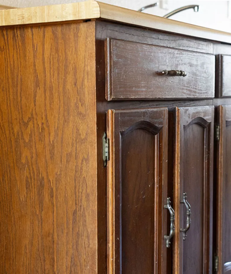 Dark wooden kitchen cabinets with aged brass handles and a light wood countertop in natural lighting.