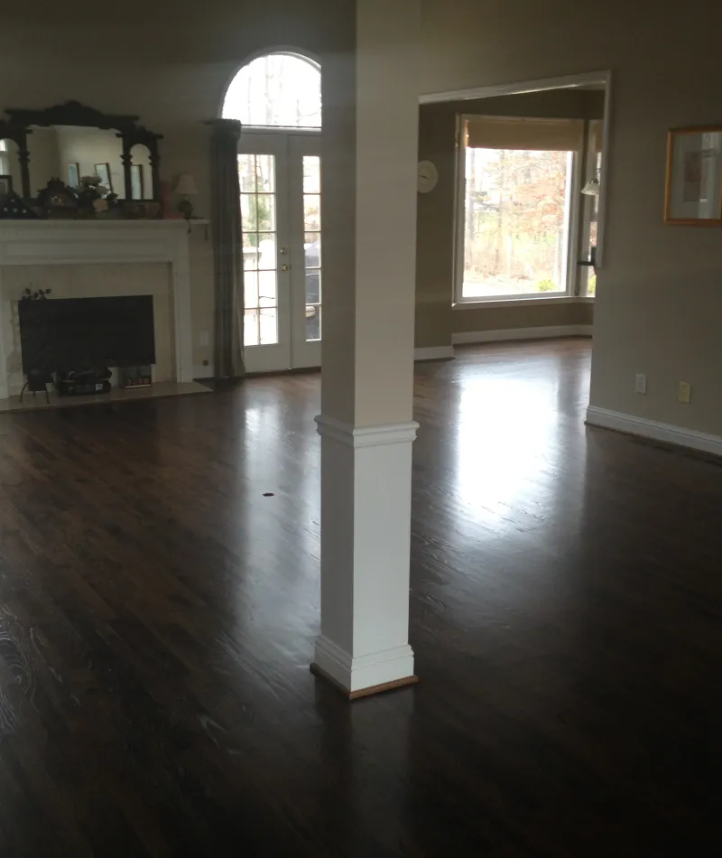 Spacious empty living room with dark hardwood floors, white fireplace, and large windows letting in natural light.