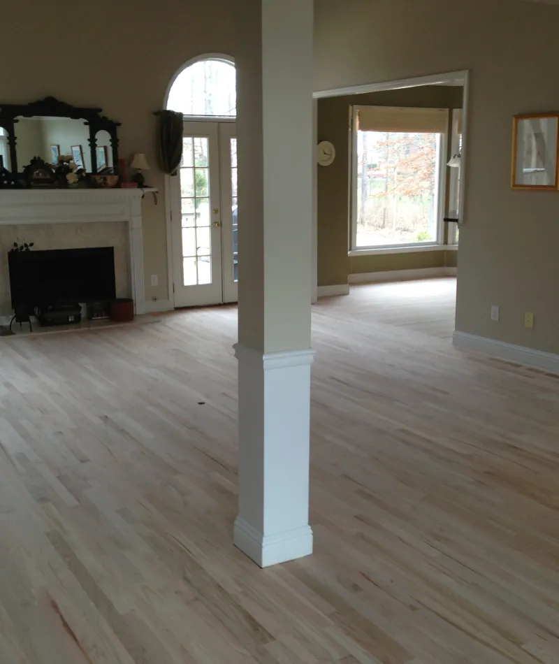 Spacious living room with light hardwood floors, white fireplace, large windows, and beige walls.