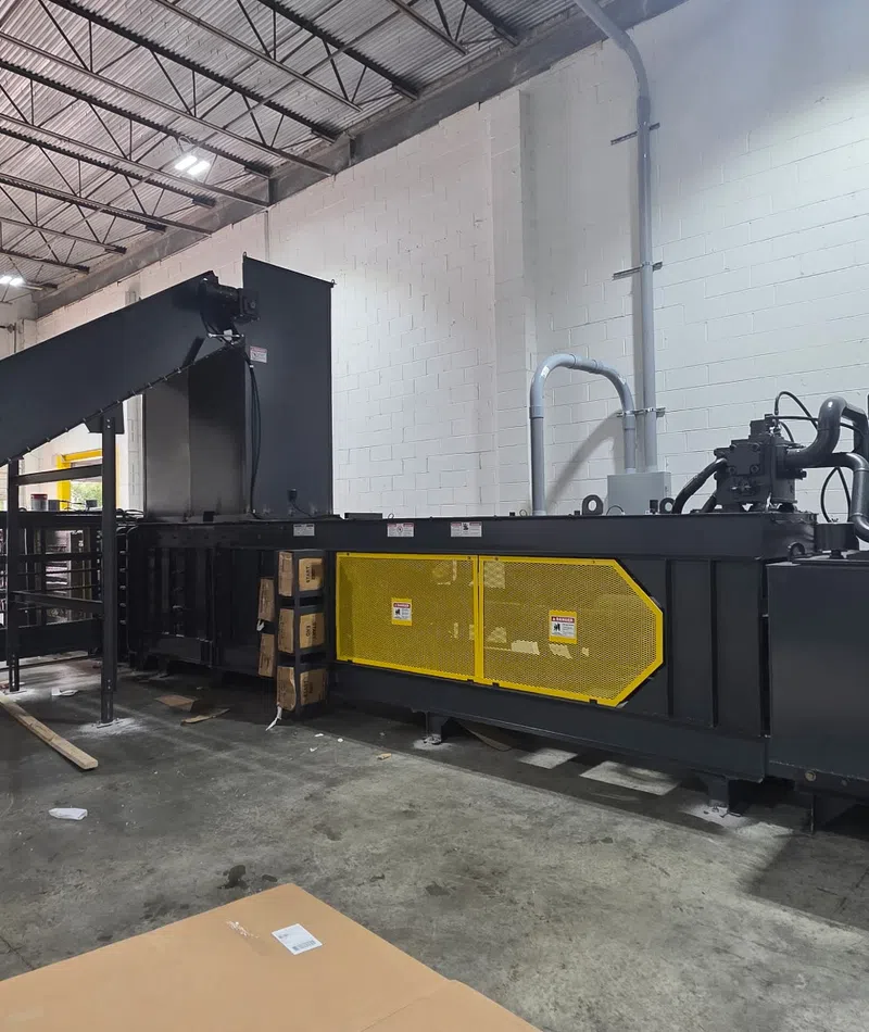 Large industrial baler machine with conveyor belt inside warehouse with concrete floor and white brick walls