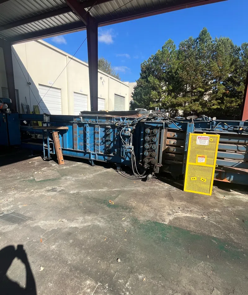 Industrial blue hydraulic baler machine with safety yellow guard, set outdoors near warehouse buildings and trees.
