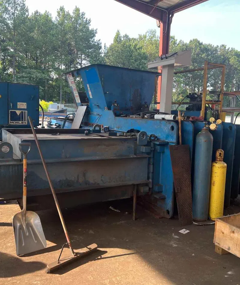Industrial metal baler machine with gas cylinders under a metal roof in an outdoor workshop area.