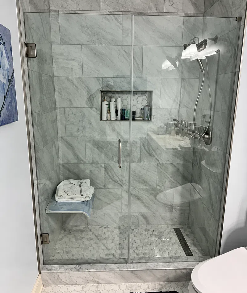 Modern bathroom shower with glass door, marble tiles, built-in shelf, and folded towels on bench.