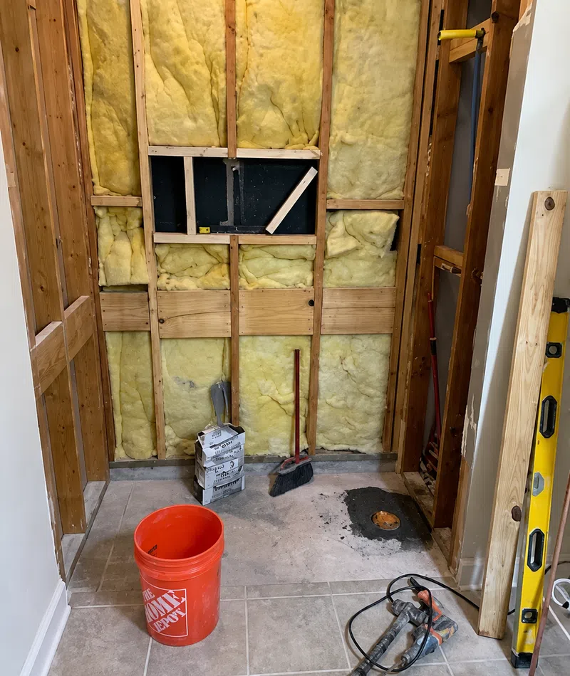 Bathroom renovation with exposed wall insulation, construction tools, red Home Depot bucket, and tiled floor.