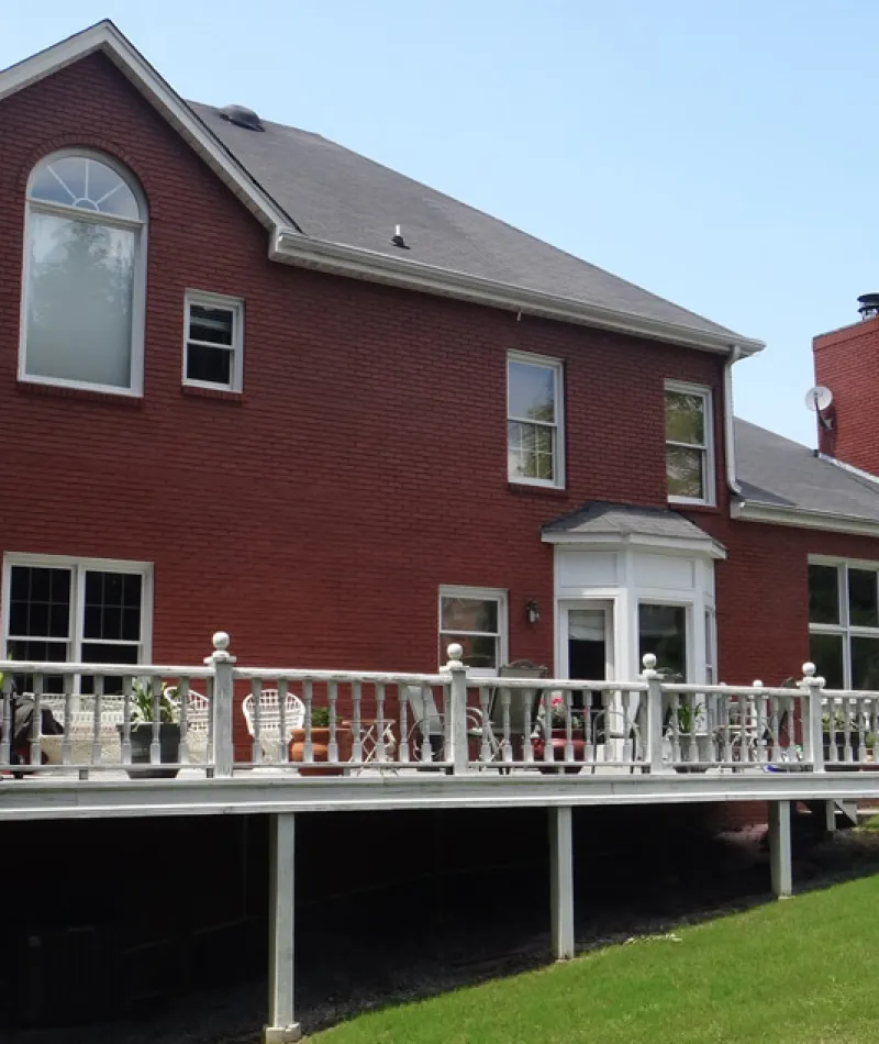 Two-story red brick house with a large elevated white deck and green lawn under clear sky.