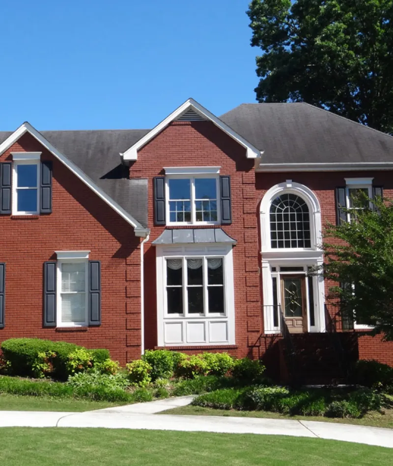 Two-story red brick house with black shutters, large arched window, and landscaped front yard under clear blue sky