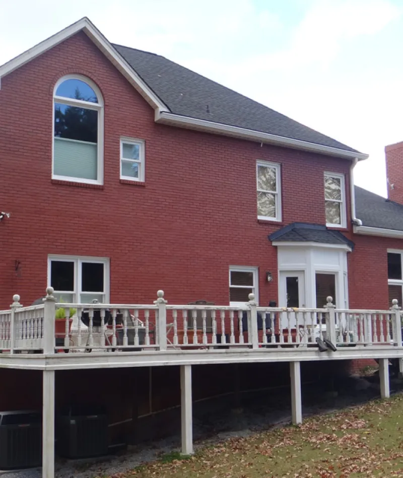 Two-story red brick house with elevated wooden deck and white railing in a suburban yard.