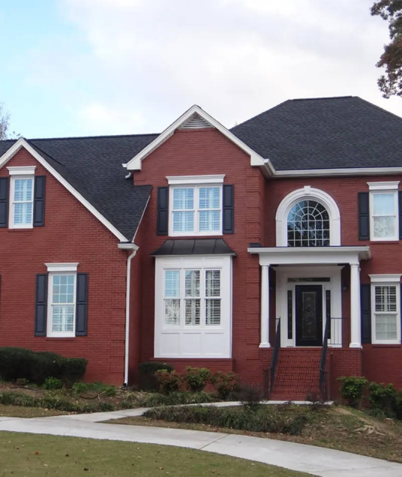 Two-story red brick house with black shutters, white trim, and a black roof surrounded by greenery and lawn.