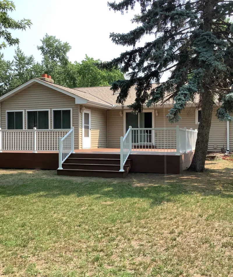 Single-story beige house with large wooden deck and white railings in spacious backyard with green lawn and trees