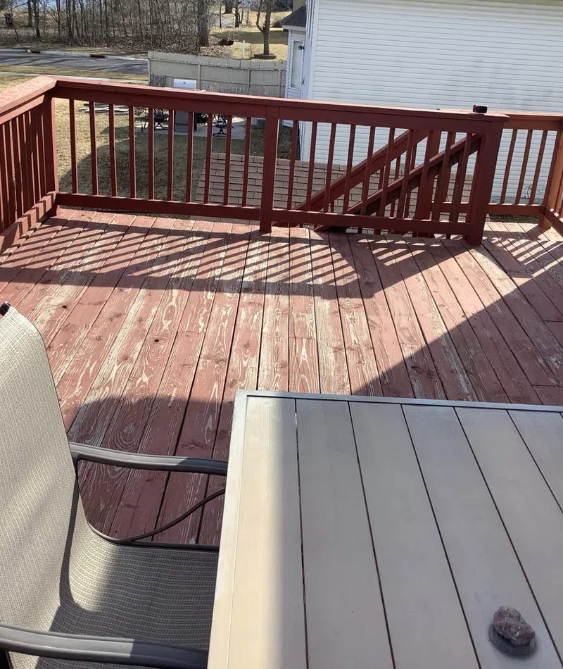 Sunny backyard deck with red wooden railings, outdoor table, and beige mesh chairs casting shadows.
