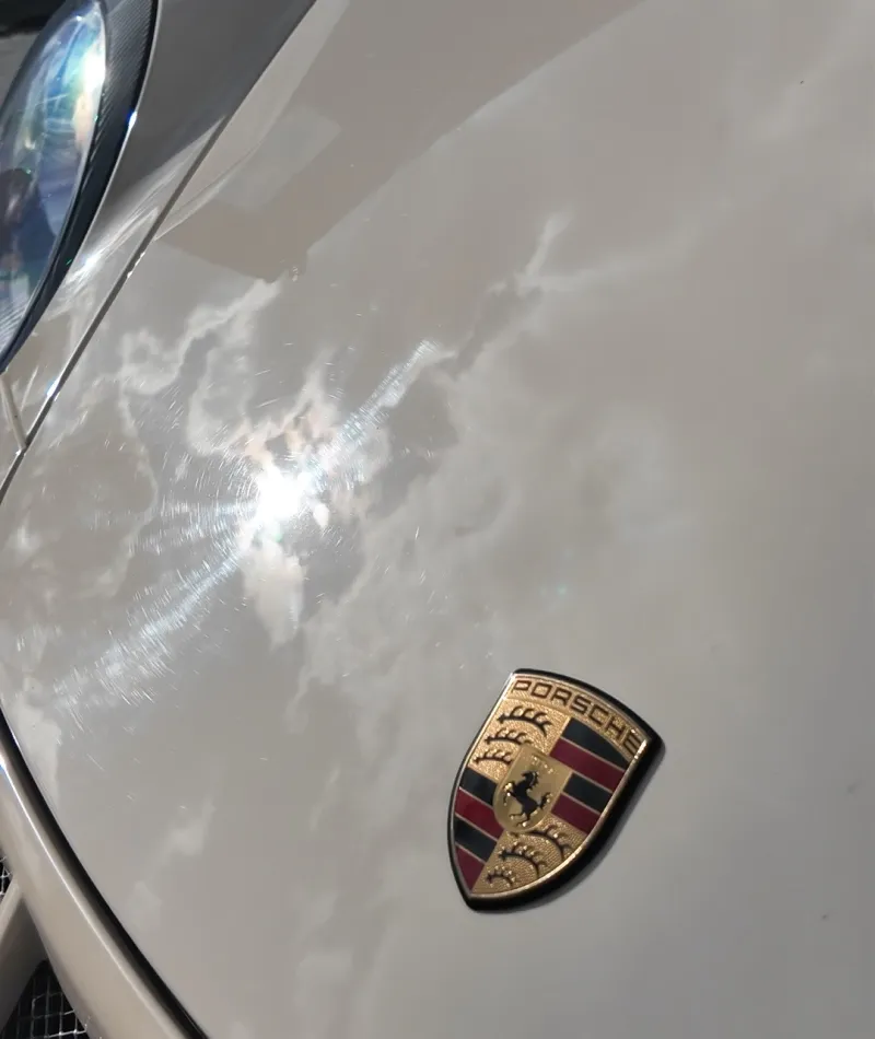 Close-up of a white Porsche car hood with emblem and reflection of clouds and sunlight