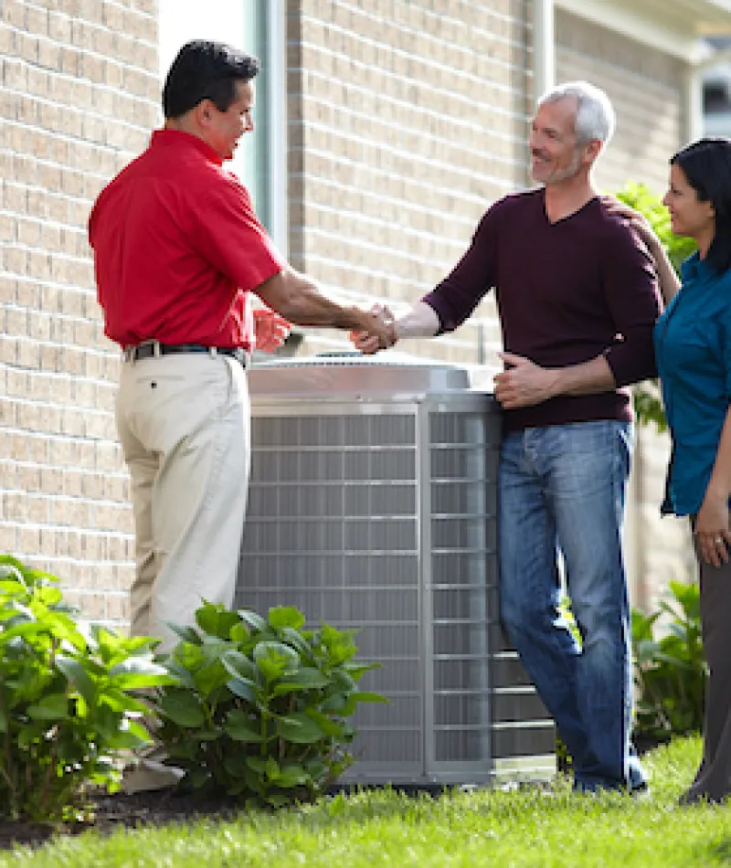 A technician shakes hands with homeowners beside a newly installed air conditioning unit.