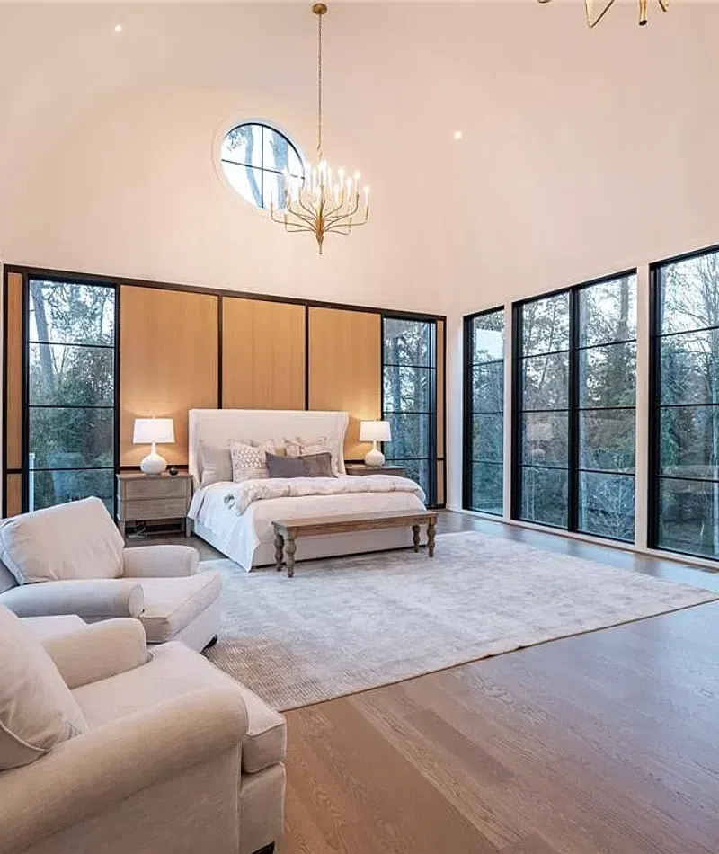 Spacious modern bedroom with large windows, high vaulted ceiling, chandelier, king bed, and two beige armchairs.