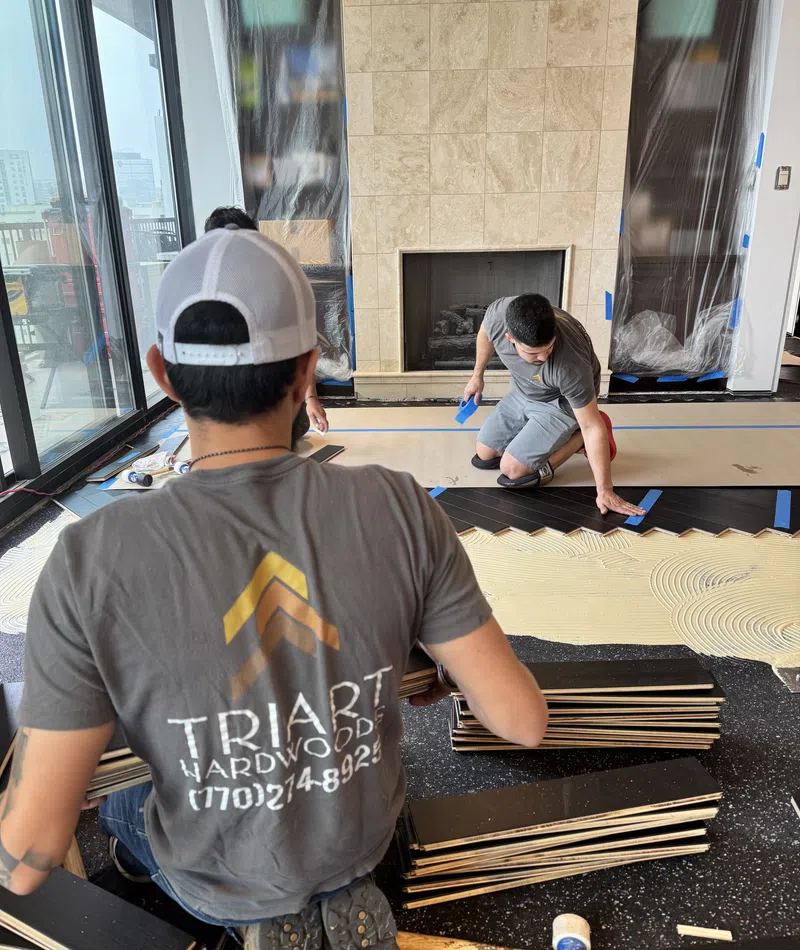 Two workers installing dark hardwood flooring in a modern living room with a fireplace and large windows.
