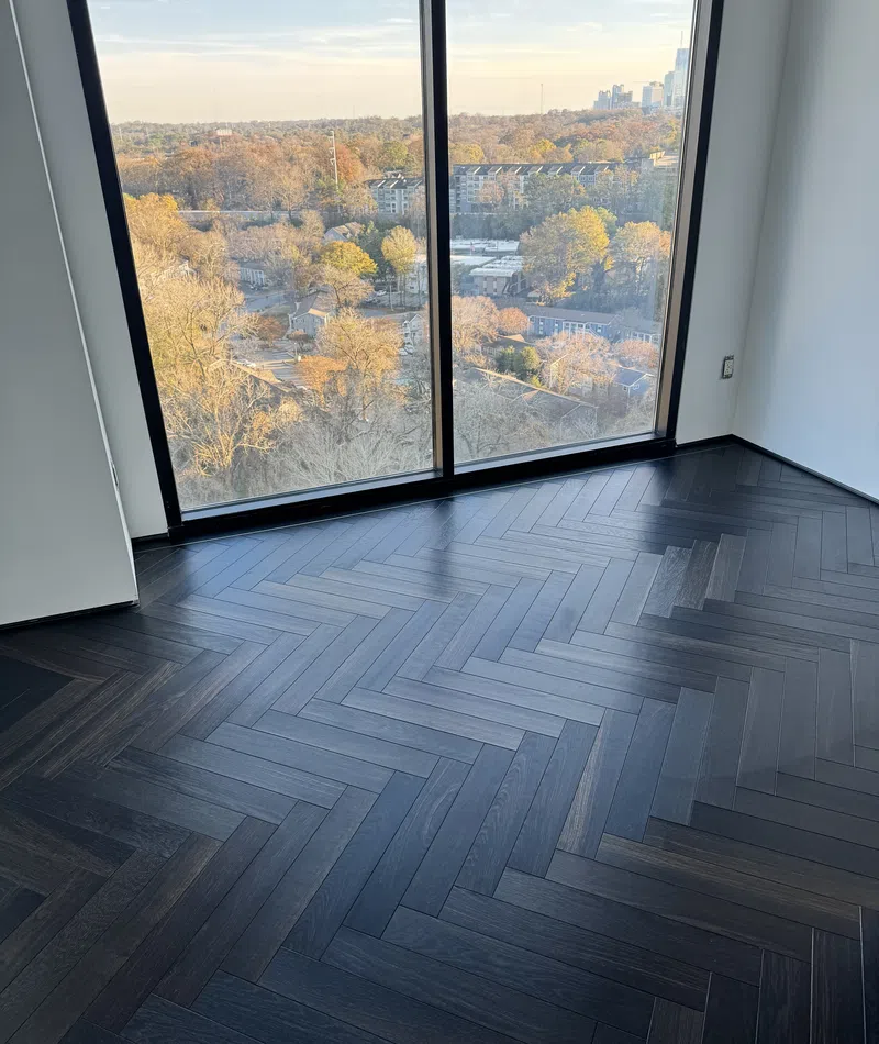 Dark herringbone wood floor in empty room with large window overlooking autumn cityscape.