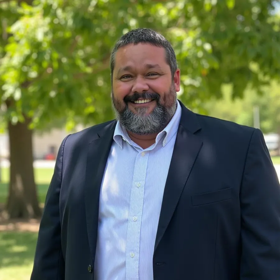 Smiling man with beard wearing a dark blazer and white shirt standing outdoors with green trees in background