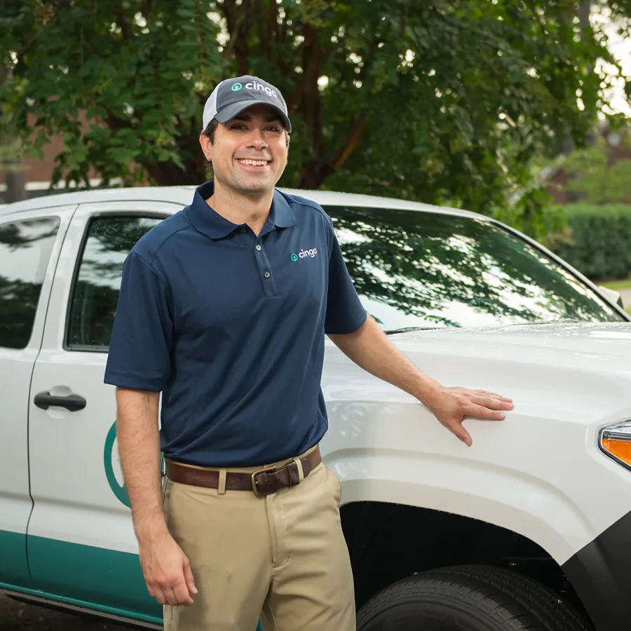 Smiling technician in uniform stands by branded white and teal service truck outdoors with greenery in background