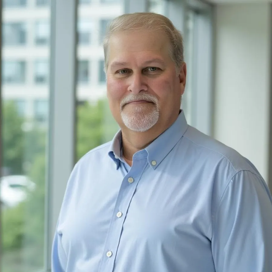 Middle-aged man with gray hair and beard in light blue shirt standing by large windows in modern office.