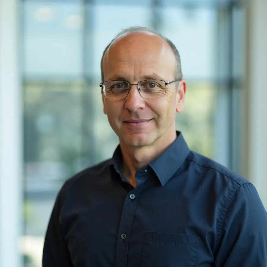 Middle-aged man with glasses and blue shirt smiling indoors by large window with blurred background